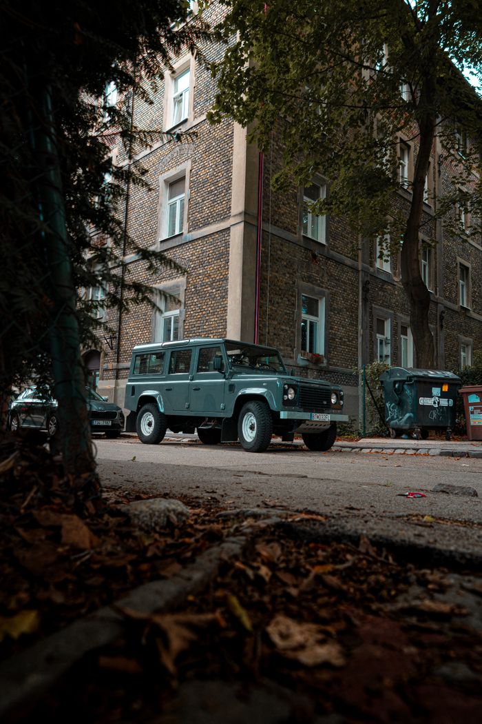 Classic defender SUV parked on city street beside a historic brick building on an autumn day.